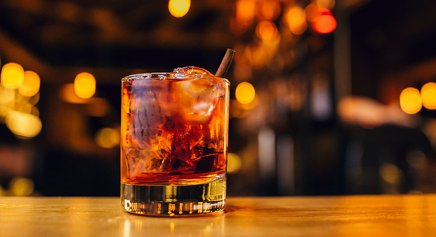 Old fashioned cocktail with a large ice cube and orange peel on a dark wooden clubhouse bar