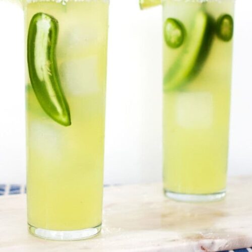 Two cocktails with jalapenos on a marble tray on a blue tablecloth.