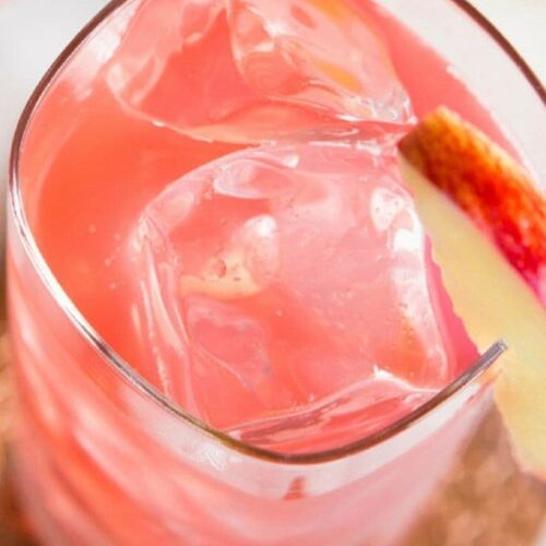 A closeup photo of a glass filled with a pink cocktail garnished with an apple.
