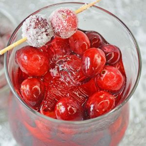 A close up of a cranberry cocktail with fresh cranberries floating in the drink with sugared cranberries on a cocktail pick as the garnish.