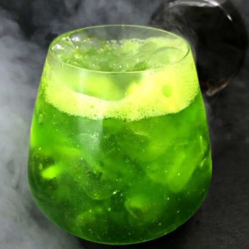 A green cocktail with bubbles and smoke on a black background with a dark potion bottle behind it.