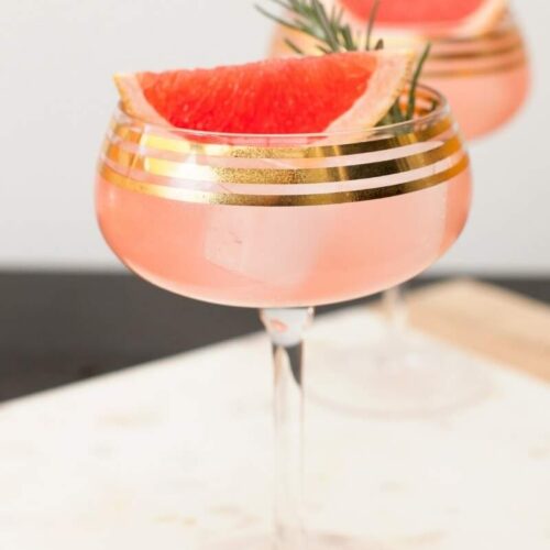 Cocktail Recipe for Elderflower Grapefruit Sparkling Martini. Served in a gold rimmed champagne coupe and garnished with a grapefruit wedge and rosemary sprig.