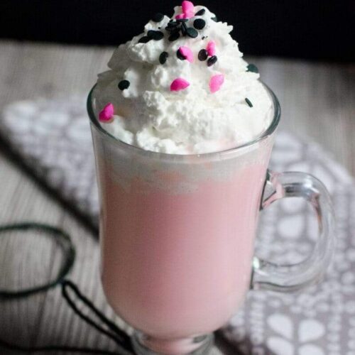 Strawberry Vanilla Hot Chocolate Recipe topped with whipped cream and sprinkles. This pink drink is so pretty served in a clear mug.