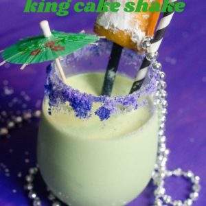 Mardi Gras King Cake Shake recipe garnished with a beignet and cocktail umbrella and strewn with mardi gras beads on a purple background.