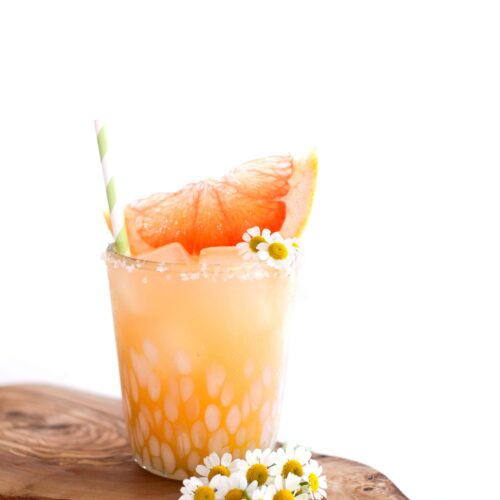 A paloma cocktail garnished with a grapefruit wedge on a wood slab with daisies.