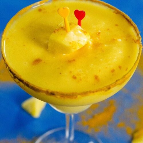 A chili mango margarita served in a chili powder rimmed margarita glass. It's on a blue background with sprinkled chili powder and chunks of mango.