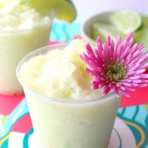A cup of Lime Dole Whip with a pink flower and lime slices as garnish on a colorful backdrop.