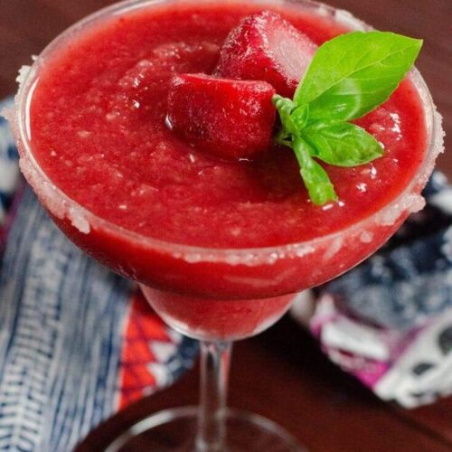 A frozen strawberry margarita garnished with fresh basil with a printed napkin on a cherry wood table.
