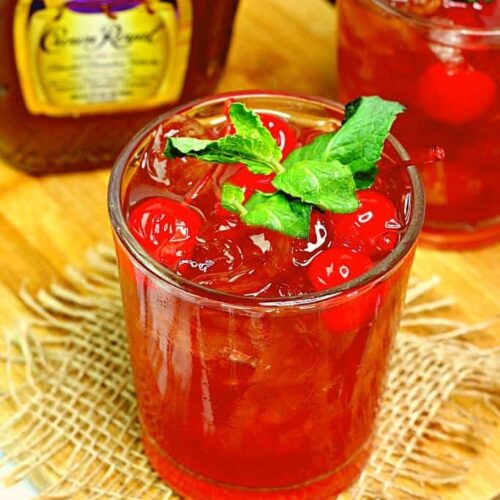 A whisky cocktail garnished with cherries and mint next to a bottle of Crown Royal whisky.