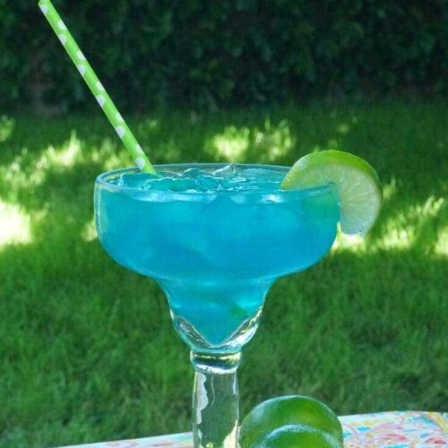 An electric blue margarita garnished with a lime wheel with a green straw on a table in a backyard