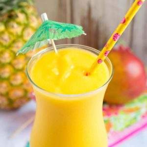 A blended cocktail with its umbrella next to a fresh pineapple and mango