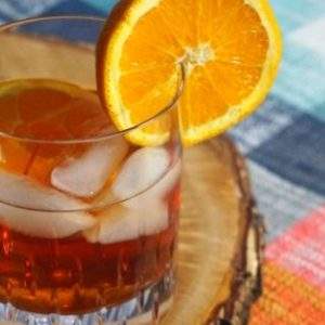 Blueberry Whiskey drink with orange wheel on a wood slice and plaid placemat