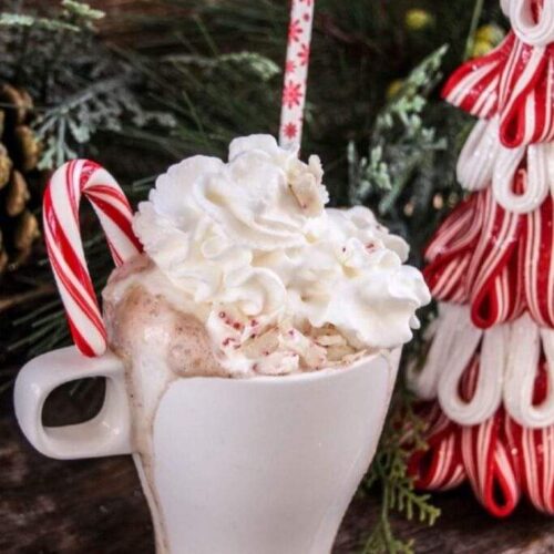 A white mug overflowing with hot chocolate topped with whipped cream and a candy cane.