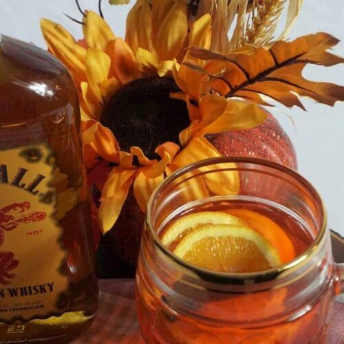 A mug of spiced cider with orange slices with a bottle of Fireball whiskey.