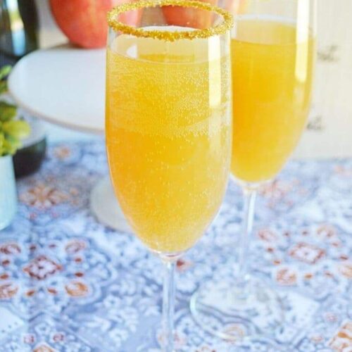 Two champagne flutes filled with apple cider mimosas on a table with a stack of apples in the background.