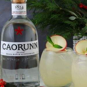 A bottle of gin next to two glasses of Caorunn holiday tea punch with red Christmas ornaments for decoration.