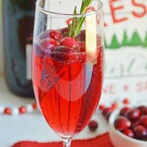 A cranberry mimosa in a champagne flute in front of a bowl of cranberries, a bottle of champagne and a Christmas sign.