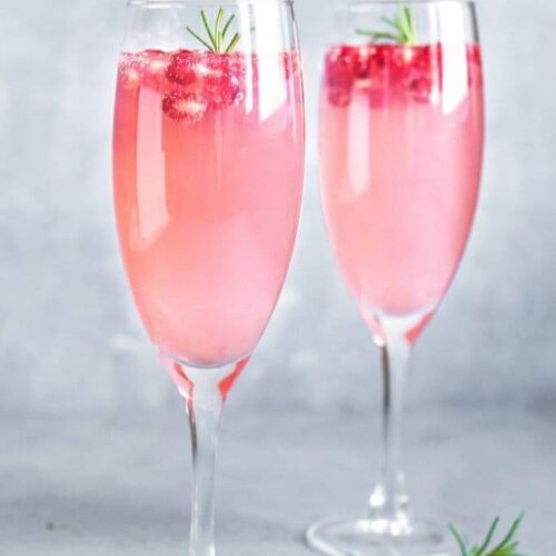 Two champagne flutes filled with French Rose Champagne Punch garnished with rosemary and pomegranate seeds on a gray backdrop.