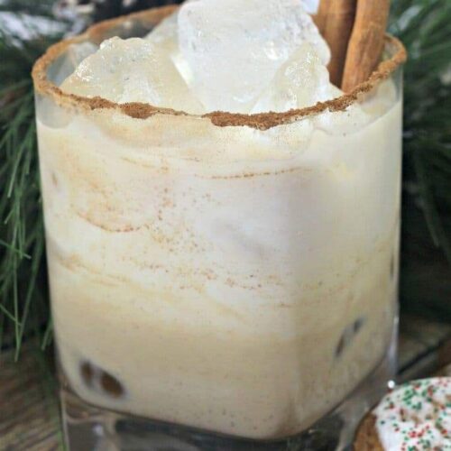 A Gingerbread White Russian cocktail garnished with cinnamon sticks next to some greenery and a gingerbread cookie.