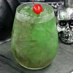 A green shimmery glass of Harry Potter inspired Death Eater's Draught cocktail on a black slate with a silver skull.