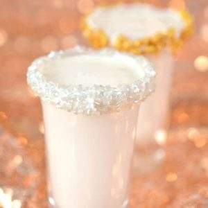 Shot glasses rimmed with silver and gold sprinkles filled with spiced rum milk shots on a rose gold sequin tablecloth.
