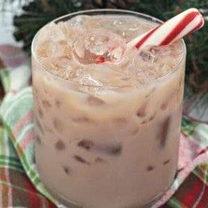 A peppermint chocolate white russian cocktail garnished with a peppermint stick on a plaid napkin next to a bottle of kahlua.