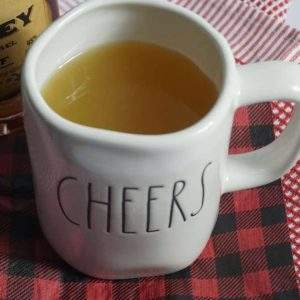 A Rae Dunn mug that says Cheers filled with Mulled Apple Cider Wassail next to a bottle of whiskey sitting on a buffalo plaid napkin.