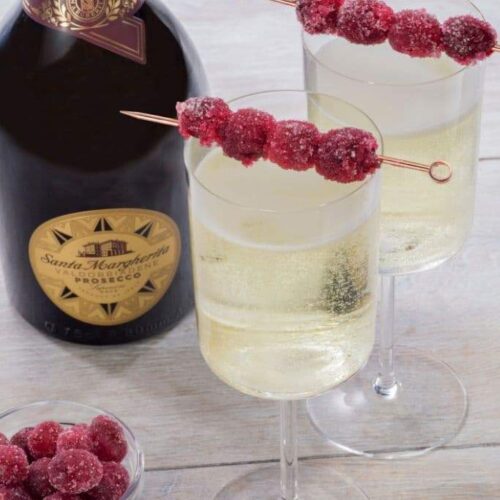 Two champagne glasses filled with Prosecco and garnished with sugared cranberries on skewers next to a bottle of prosecco and a bowl of cranberries.