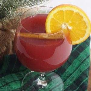 A glass of slow cooker hot holiday punch garnished with a cinnamon stick and orange slice on a green plaid napkin next to a small Christmas tree.