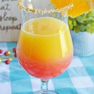 A sunrise mimosa cocktail in a small hurricane glass garnished with an orange wedge and sprinkles on a blue gingham tablecloth.