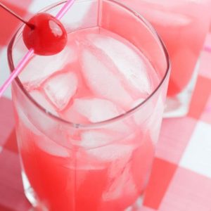 A marashchino cherry on a skewer rests on a glass of pink lemonade on a red and white gingham tablecloth.