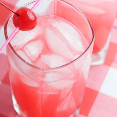 A marashchino cherry on a skewer rests on a glass of pink lemonade on a red and white gingham tablecloth.