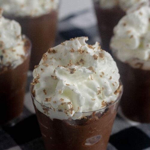 Chocolate pudding shots topped with whipped cream on a black and white tablecloth.
