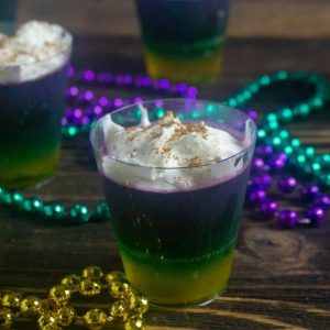 Three colored Jello shots decorated for Mardi Gras on a wood table.