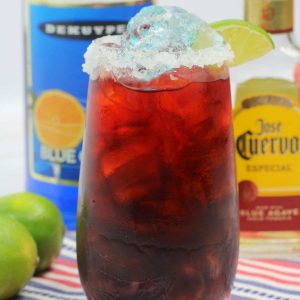 A red cocktail garnished with lime with a bottle of blue curacao and Jose Cuervo tequila.
