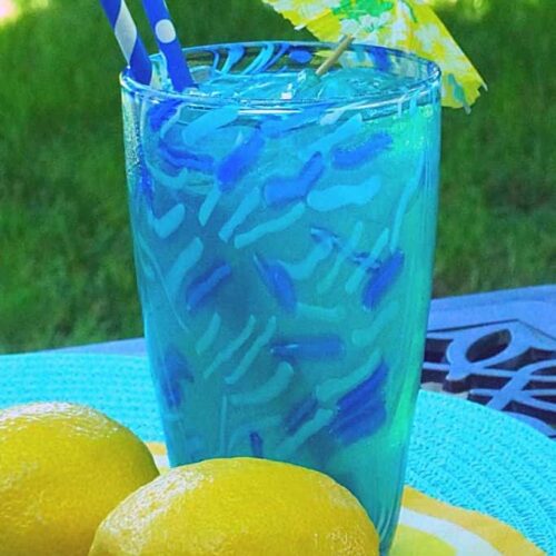 A glass of blue lemonade with an umbrella and two paper straws on a table next to lemons.