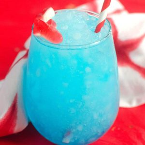 A red gummy shark sits on the edge of a glass filled with a blue slushie on a red table.