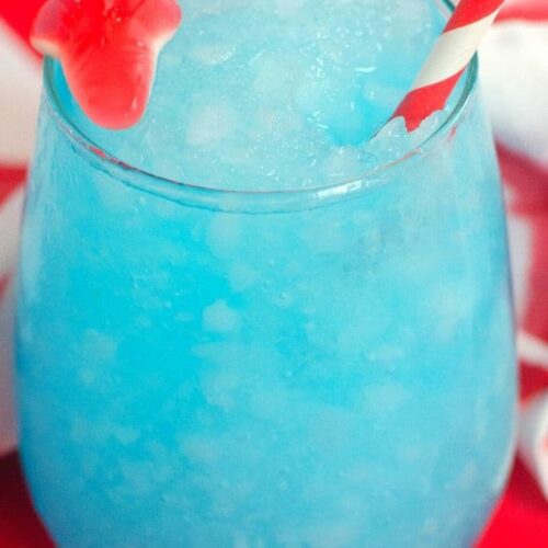 A blue frozen drink with a red gummy shark and paper straw on a red table.