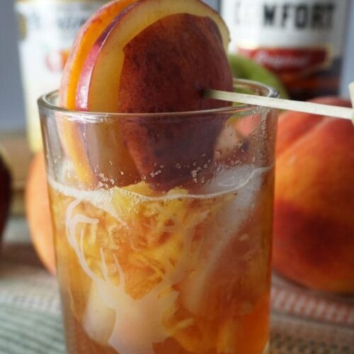 A cocktail garnished with fresh peaches and apples with fruit and a bottle of cider and Southern Comfort.