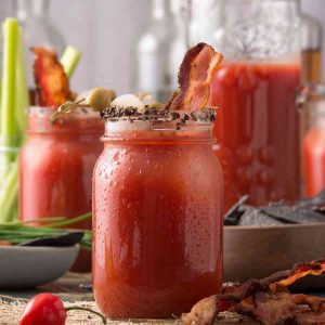 A mason jar filled with clamato juice with bacon slices, and a cocktail olive garnish with other beverages in the background.