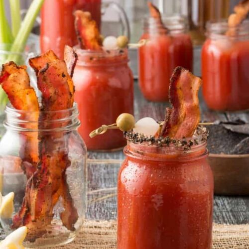 Mason jars filled with tomato cocktail garnished with bacon on a table.