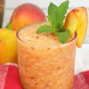 A frozen margarita garnished with a peach slice and mint next to some fresh peaches.