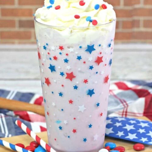 A milkshake topped with whipped cream and sprinkles in a glass decorated with red, white, and blue stars. It's on a tray with USA M&Ms and paper straws.