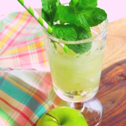 A green slushie topped with mint on a wood tray with a plaid napkin and a green apple.