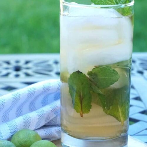 A cocktail with mint leaves rests on a wood plate on a patio table.