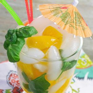 A cocktail with fresh basil and peaches with an orange umbrella.