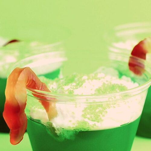 Plastic cups filled with lime Jello, whipped cream and topped with gummy worms on a green background.