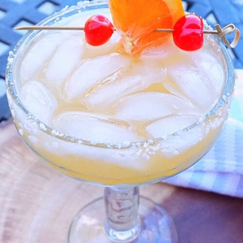 A margarita garnished with an orange wheel and maraschino cherries on a patio table.