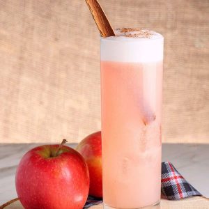 An apple next to a pink cocktail topped with foam and a cinnamon stick on a plaid napkin.