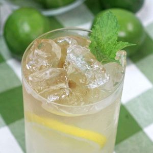 A glass of lemonade with with a slice of lemon floating in the drink next to a pile of limes.